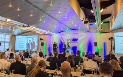 Zwei Moderatoren stehen auf der Bühne während des POSY OutputForum 2024 mit Publikum im Hintergrund.